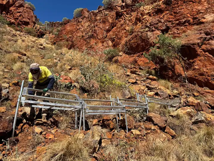 Field worker carrying a metal scaffold structure across red-rock terrain for a remote installation.