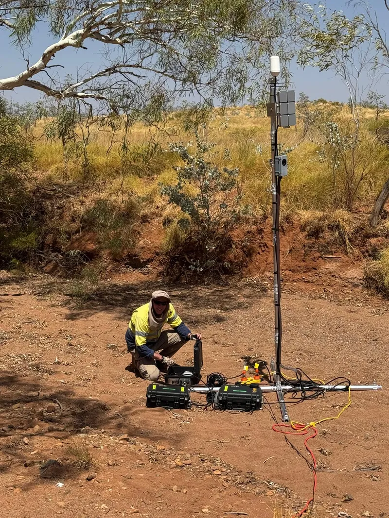 Field engineer setting up an outdoor sensor mast with battery boxes and cabling at an Australian outback site.